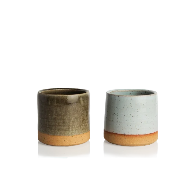 Two small ceramic cups side by side, one glazed in olive green, one in speckled pale blue, both with unglazed terracotta bases, resting on a warm neutral surface in soft daylight.